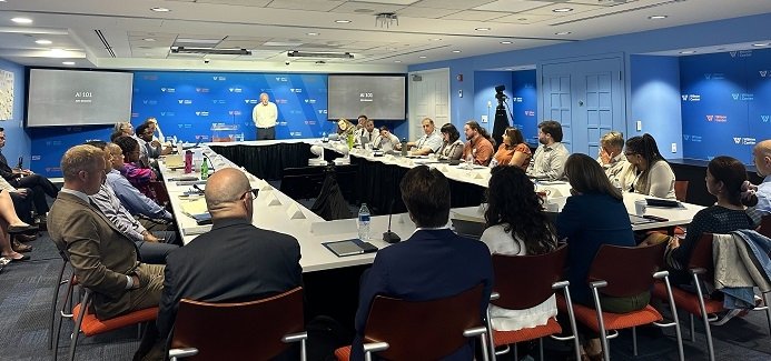 Room filled with people seated at a table with a speaker at the front, on the screens it says AI 101