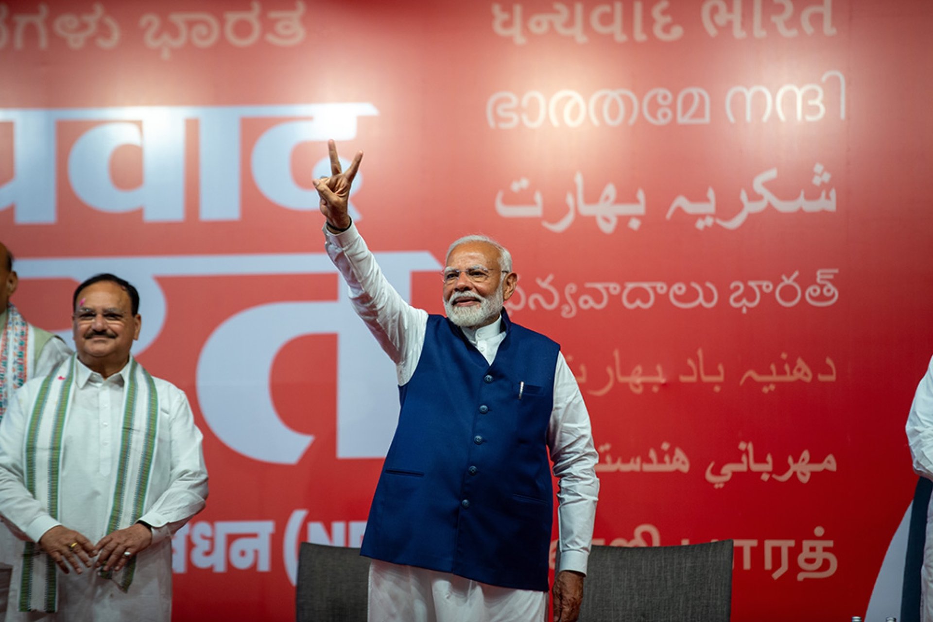 Narendra Modi stands in front of a red background, holding up his hand in a victory sign.