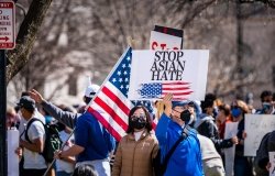 A man stands in a group of protestors wearing a masks, holding a sign with an American flag on it that says Stop Asian Hate.